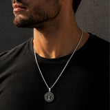 A close-up of a man wearing a silver chain necklace with a circular pendant featuring a tree of life design, set against a dark gray background.