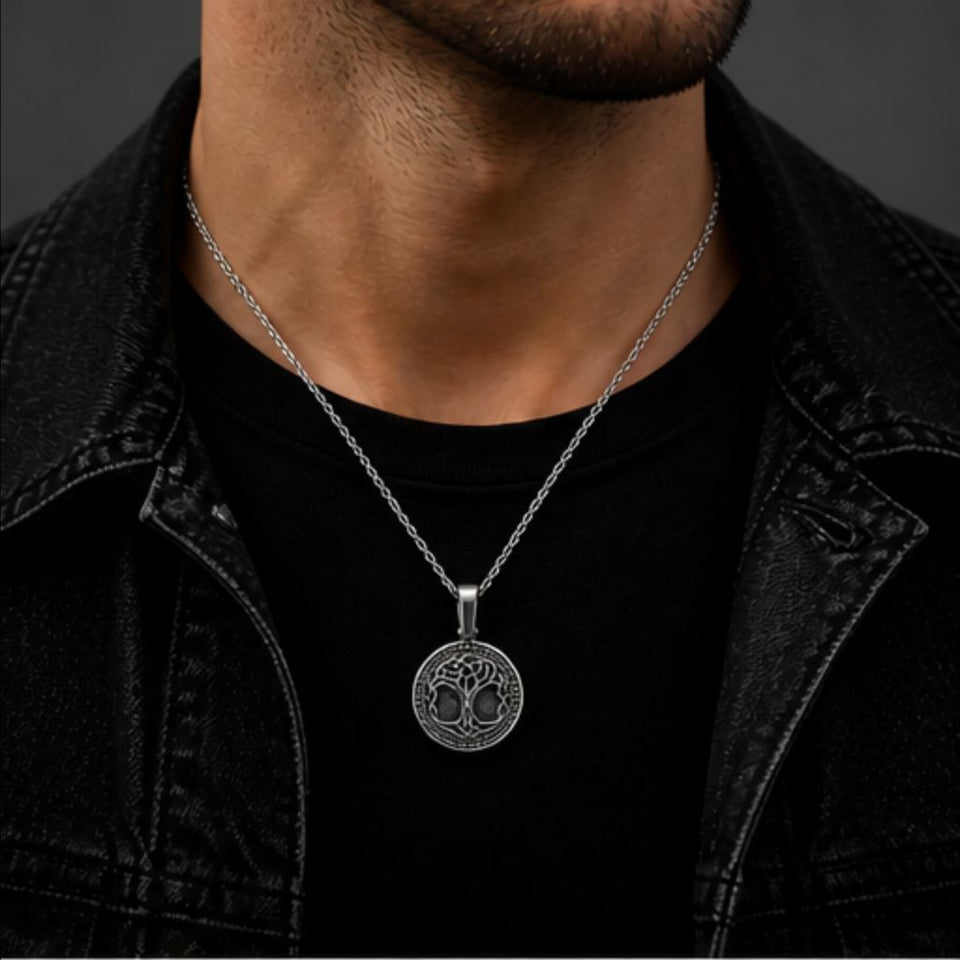 A silver pendant necklace with a detailed circular emblem, worn by a man over a black t-shirt and black denim jacket.