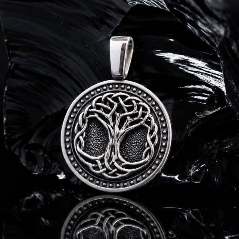 Handcrafted 925 sterling silver Tree of Life pendant with oxidized details. A silver pendant featuring an intricate Celtic knot tree design, displayed on a reflective black surface with a dark, textured rock in the background.