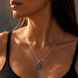 A close-up lifestyle shot of a woman wearing a silver chain necklace with a circular pendant featuring a tree of life design, paired with a black ribbed tank top.
