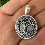 Detailed close-up of Norse Yggdrasil Tree of Life engraved silver necklace. A silver pendant with an intricate Celtic knot tree design, held in a person's hand to show scale.